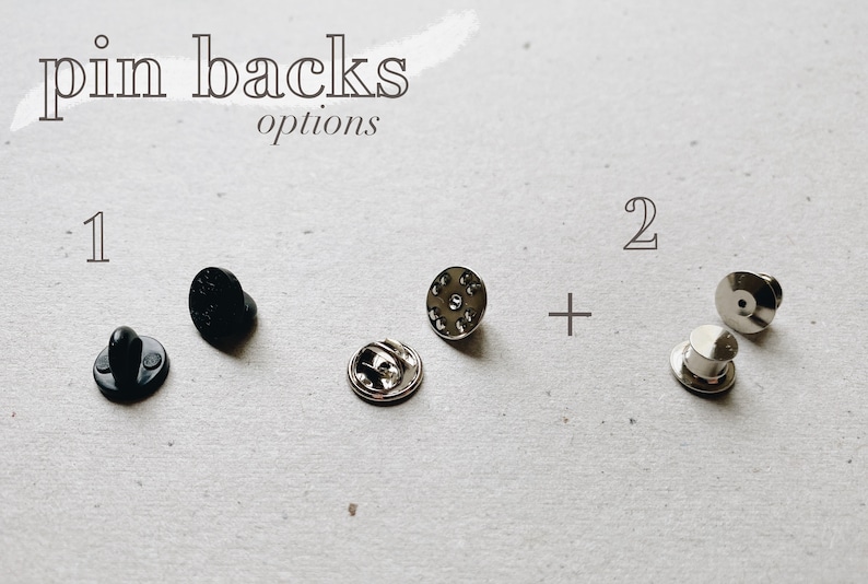 May include: Two different types of pin backs. The first type is black and has a post on the back. The second type is silver and has a post on the back. The text "pin backs options" is written above the pin backs. The number "1" is written above the black pin backs and the number "2" is written above the silver pin backs.