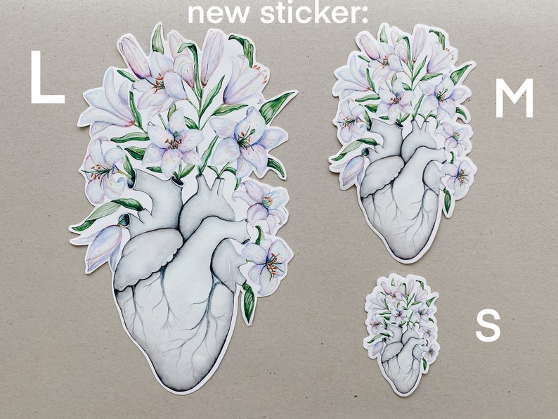 May include: Three sizes of a sticker featuring a white anatomical heart with white lilies growing from the top. The sticker is available in sizes L, M, and S.