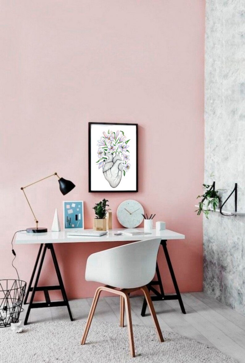 May include: A pink wall with a white desk and a white chair. A framed print of a heart with flowers is hanging on the wall. The desk has a lamp, a clock, and a plant on it.
