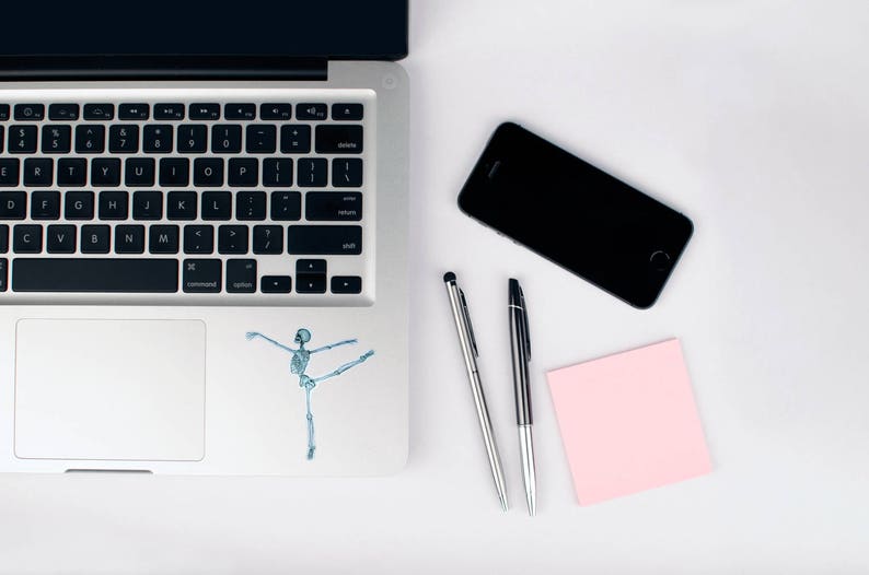 May include: A silver laptop computer with a black keyboard and a white trackpad. A skeleton sticker is on the trackpad. A black smartphone, two silver pens, and a pink sticky note are on a white surface.