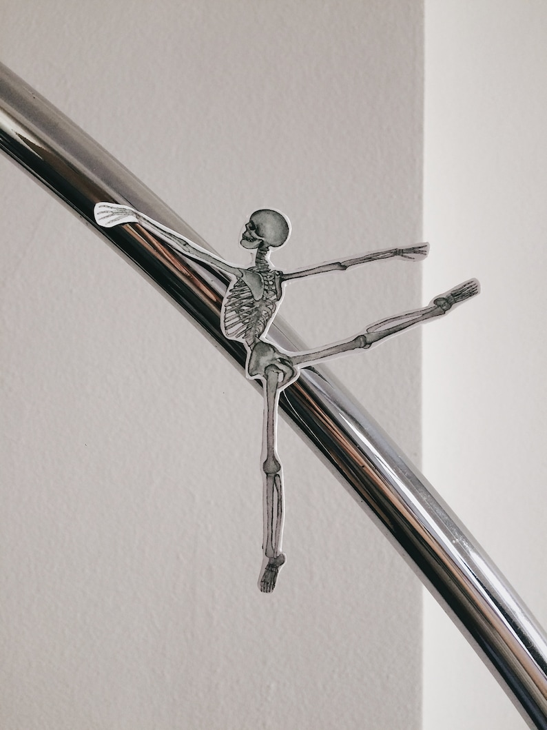 May include: A black and white illustration of a skeleton in a ballet pose, with one leg extended and arms outstretched. The skeleton is positioned against a shiny, silver metal bar, with a white wall in the background.