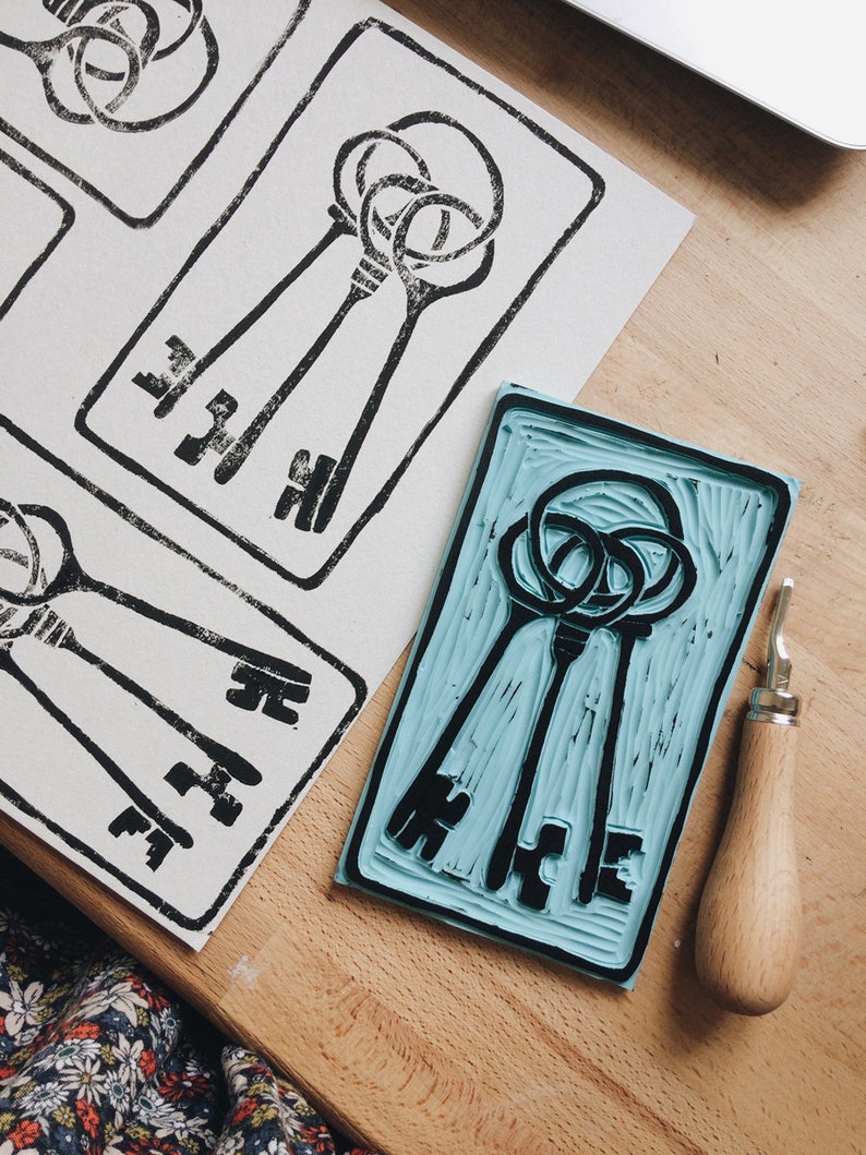 Może przedstawiać: Zbliżenie na niebieską gumową pieczątkę z trzema wyrzeźbionymi czarnymi kluczami. Pieczątka leży na drewnianej powierzchni obok drewnianego narzędzia do rzeźbienia.