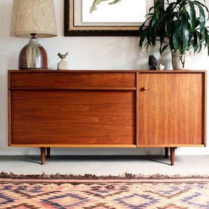 May include: A vintage-style wooden sideboard with a single drawer and a cabinet door. The sideboard is on a patterned rug with a geometric design in shades of pink, blue, and brown. A framed print of a whale skeleton is hanging on the wall above the sideboard.