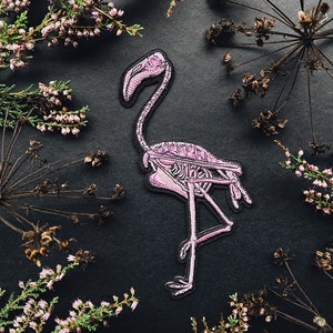 K&ouml;nnte beinhalten: Rosa bestickter Flamingo-Skelett-Patch auf schwarzem Hintergrund, umgeben von getrockneten Blumen.