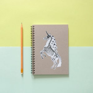 May include: A brown notebook with a spiral binding and a black and white sticker of a unicorn skeleton on the front.