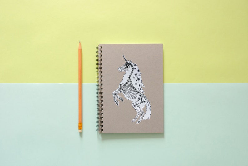 May include: A brown notebook with a spiral binding and a black and white sticker of a unicorn skeleton on the front.