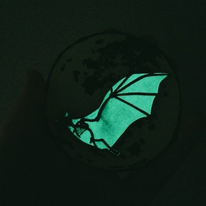 Puede incluir: Una pegatina que brilla en la oscuridad de un murci&eacute;lago con las alas extendidas, volando frente a una luna.