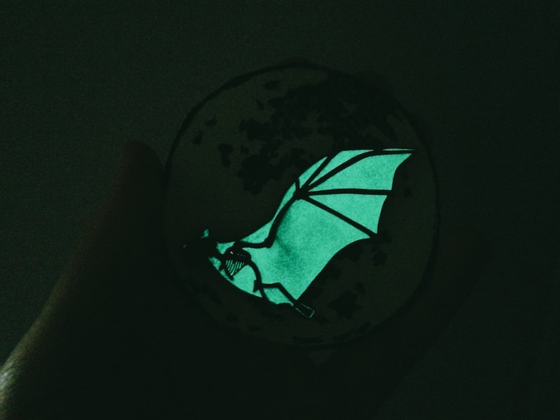 Puede incluir: Una pegatina que brilla en la oscuridad de un murci&eacute;lago con las alas extendidas, volando frente a una luna.