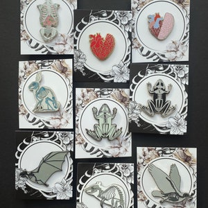 Puede incluir: Conjunto de 10 pines de esmalte con ilustraciones anat&oacute;micas de varios animales y &oacute;rganos. Los pines est&aacute;n en tarjetas blancas con dise&ntilde;os florales en blanco y negro. Los pines incluyen un coraz&oacute;n, un cerebro, una rana, un murci&eacute;lago, una rata, un conejo y un esqueleto humano.