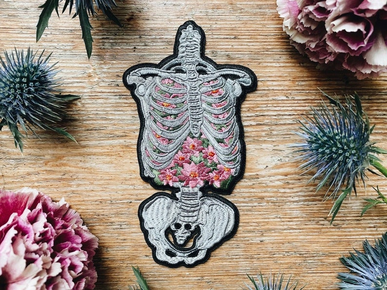 May include: Embroidered patch featuring a skeletal torso filled with pink flowers. The patch has a black border and is set against a wooden background, with decorative flowers and thistles surrounding it. The patch is a unique, gothic-inspired design.