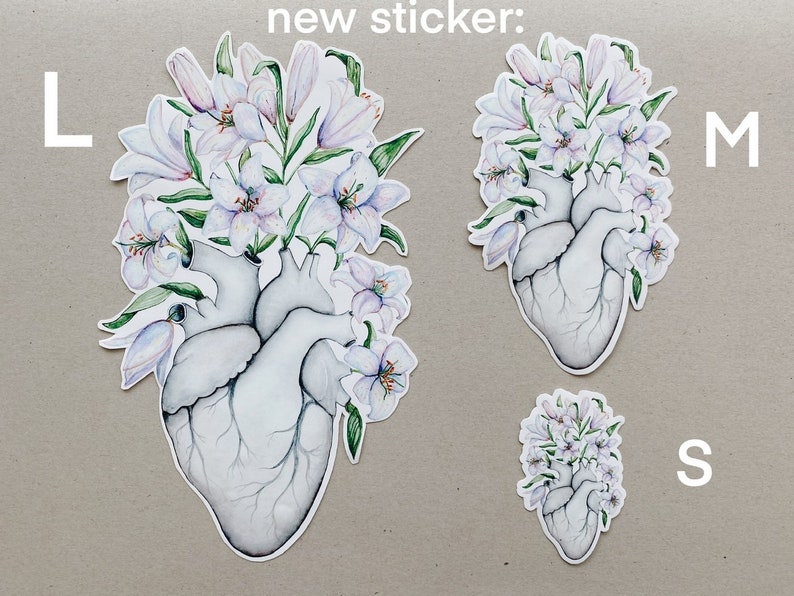 May include: Three sizes of a sticker featuring a white anatomical heart with white lilies growing from it. The sizes are labeled "L", "M", and "S".