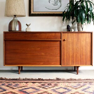May include: A mid-century modern wooden cabinet with a single drawer and a door. The cabinet is on a rug with a geometric pattern in shades of pink, blue, and brown. A lamp with a beige shade and a ceramic base sits on top of the cabinet. A framed print of a rat skeleton is on the wall above the cabinet.