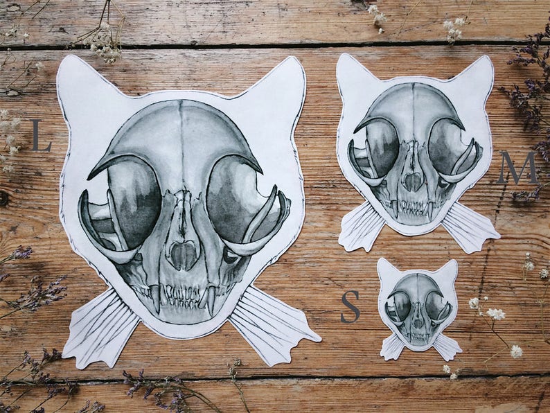 May include: Three black and white cat skull illustrations on a wooden background. The illustrations are in different sizes, with the largest one being labelled "L", the medium one labelled "M", and the smallest one labelled "S".