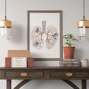 May include: A framed illustration of a stylized human lung with pink flowers and blue birds. The illustration is hanging on a white wall above a wooden console table with two drawers. There are two gold pendant lights hanging above the table, and a potted plant, books, and a candle on the table.