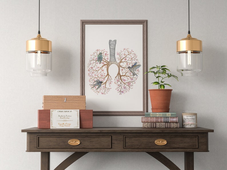 May include: A framed illustration of a stylized human lung with pink flowers and blue birds. The illustration is hanging on a white wall above a wooden console table with two drawers. There are two gold pendant lights hanging above the table, and a potted plant, books, and a candle on the table.