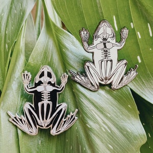 Op de afbeelding: Twee zilveren kikker skelet pinnen op een groene bladeren achtergrond. De kikker skeletten zijn gedetailleerd en hebben een zwarte omtrek.