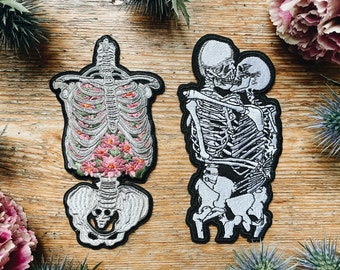 Könnte beinhalten: Zwei gestickte Bügelbilder mit Skeletten. Ein Patch zeigt ein Skelett mit einem Blumendesign im Brustkorb. Der andere Patch zeigt zwei Skelette, die sich umarmen.