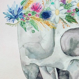 以下が含まれることがあります： 水彩画で描かれた人間の頭蓋骨に花冠が飾られています。花冠は青、白、ピンク、黄色の花と緑の葉、小麦の穂でできています。