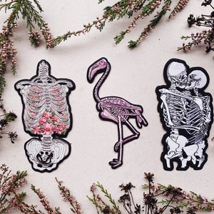 K&ouml;nnte beinhalten: Drei gestickte B&uuml;gelbilder mit Skeletten. Ein Patch zeigt einen Skelett-Torso mit rosa Blumen, ein anderer zeigt ein Flamingo-Skelett und der letzte zeigt zwei Skelette, die sich umarmen.