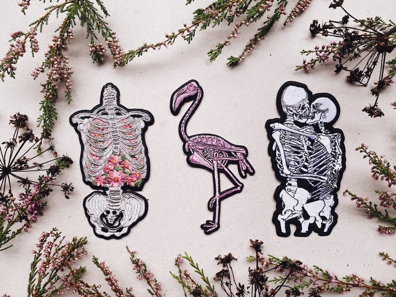K&ouml;nnte beinhalten: Drei gestickte B&uuml;gelbilder mit Skeletten. Ein Patch zeigt einen Skelett-Torso mit rosa Blumen, ein anderer zeigt ein Flamingo-Skelett und der letzte zeigt zwei Skelette, die sich umarmen.