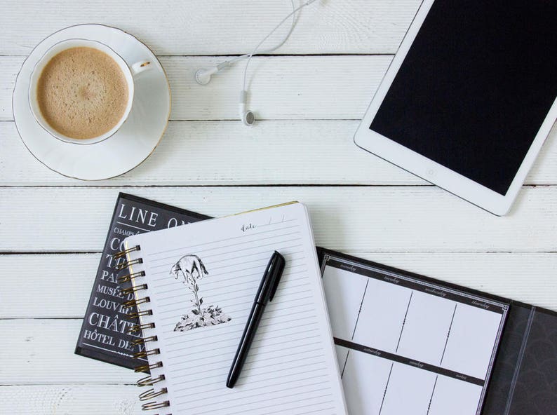 May include: A top-down view of a workspace with a cup of coffee, a tablet, and a notebook with a pen. The notebook has a sketch of a tree. White earbuds are next to the tablet. The items are on a white wooden surface.