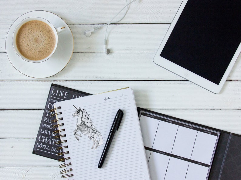May include: A white desk with a cup of coffee, a notebook with a pen and a unicorn drawing, a black planner, and white earbuds.