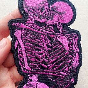 K&ouml;nnte beinhalten: Rosa gestickter Patch mit zwei sich umarmenden Skeletten. Die Skelette sind schwarz umrandet und haben ein rosa gesticktes Design.