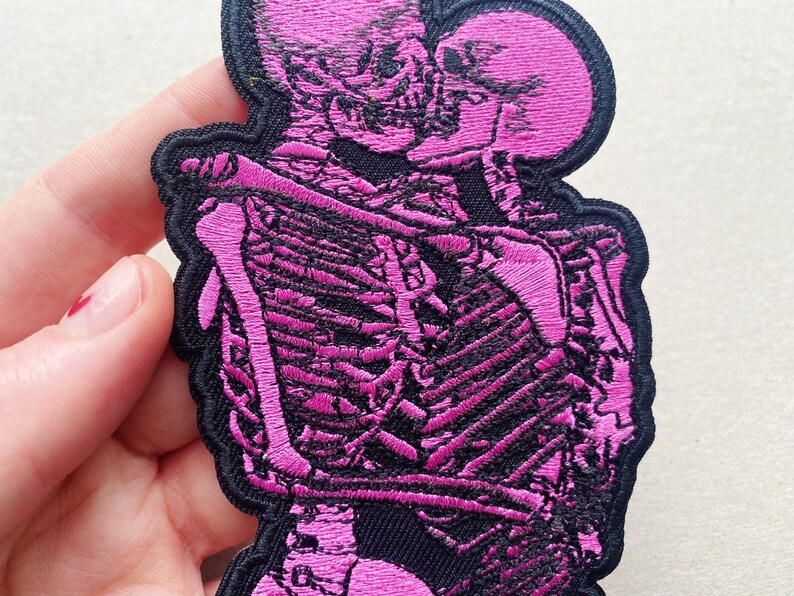 K&ouml;nnte beinhalten: Rosa gestickter Patch mit zwei sich umarmenden Skeletten. Die Skelette sind schwarz umrandet und haben ein rosa gesticktes Design.