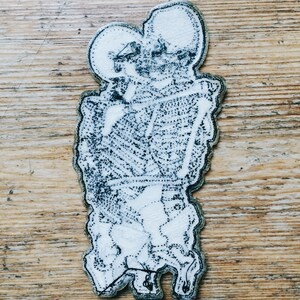 May include: A white embroidered patch with a black outline depicting a cartoon-like figure with two heads and a body that resembles a human figure. The figure is holding a rectangular object in its hands.