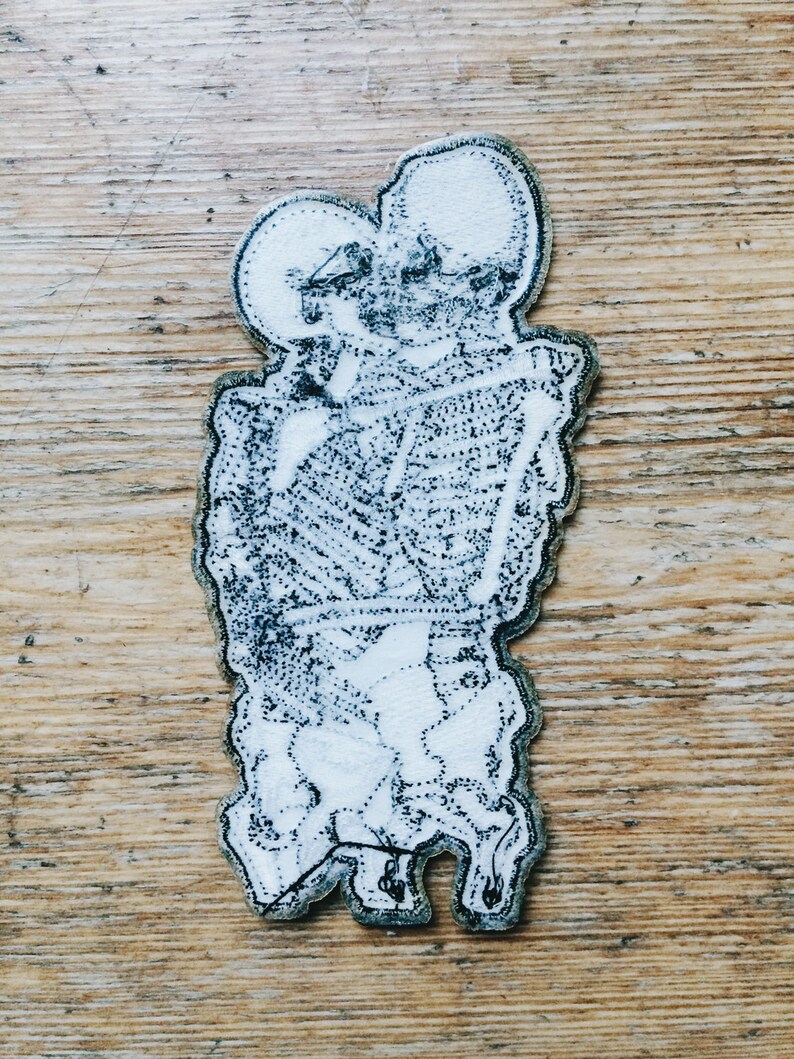 May include: A white embroidered patch with a black outline depicting a cartoon-like figure with two heads and a body that resembles a human figure. The figure is holding a rectangular object in its hands.