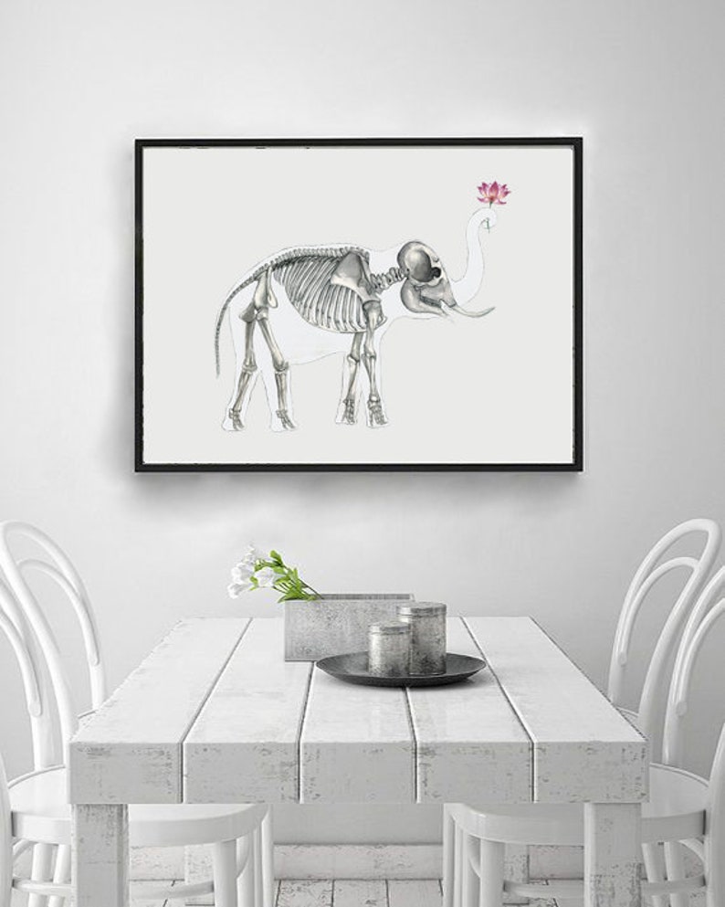 May include: A black framed print of a white elephant skeleton with a pink lotus flower above its head. The print is hanging on a white wall above a white table with two white chairs.