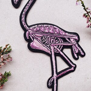 K&ouml;nnte beinhalten: Rosa-farbener Flamingo-Skelett-Stickerei-Aufn&auml;her mit schwarzen N&auml;hten. Der Flamingo steht auf einem Bein und hat den Kopf zur Seite geneigt.