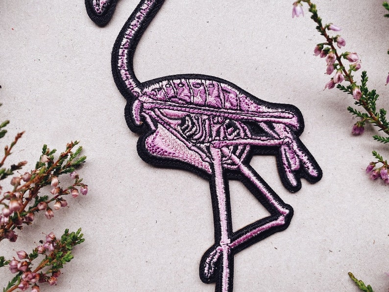 K&ouml;nnte beinhalten: Rosa-farbener Flamingo-Skelett-Stickerei-Aufn&auml;her mit schwarzen N&auml;hten. Der Flamingo steht auf einem Bein und hat den Kopf zur Seite geneigt.
