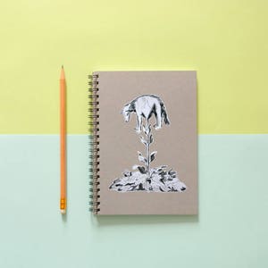 May include: A spiral-bound notebook with a brown cover featuring a black and white illustration of a wolf atop a plant. A yellow pencil is positioned to the left of the notebook. The background is split between light green and yellow.