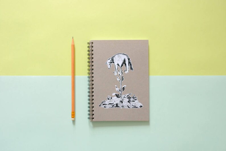 May include: A spiral-bound notebook with a brown cover featuring a black and white illustration of a wolf atop a plant. A yellow pencil is positioned to the left of the notebook. The background is split between light green and yellow.