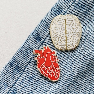 以下が含まれることがあります： 青いデニムの背景に2つのエナメルピン。1つのピンは白い脳のアウトライン、もう1つは赤い心臓のアウトラインです。