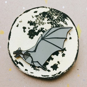 Puede incluir: Un pin de esmalte plateado de un esqueleto de murci&eacute;lago con las alas extendidas, sobre un fondo blanco con salpicaduras de pintura negra, que se asemeja a la luna.