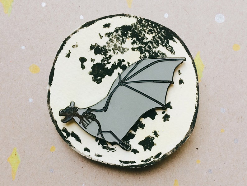 Puede incluir: Un pin de esmalte plateado de un esqueleto de murci&eacute;lago con las alas extendidas, sobre un fondo blanco con salpicaduras de pintura negra, que se asemeja a la luna.