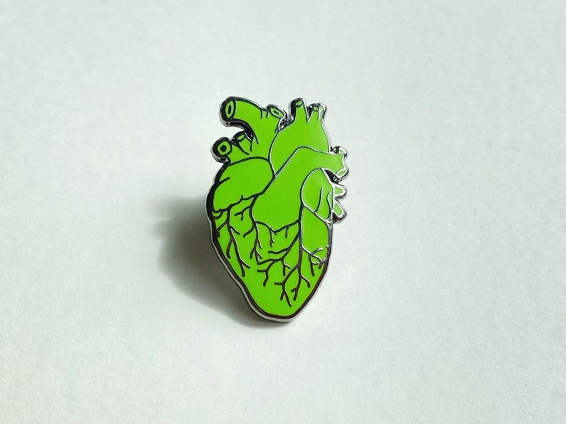 May include: A green anatomical heart-shaped enamel pin with black vein detailing and a silver border. The pin is a stylized representation of a human heart, suitable for medical professionals or those with an interest in anatomy.