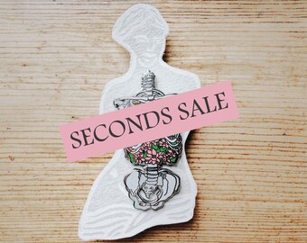 Könnte beinhalten: Eine weiße Umrandung eines menschlichen Skeletts mit rosa Blumen im Brustkorb. Das Bild hat ein rosa Banner darüber, auf dem "SECONDS SALE" steht.
