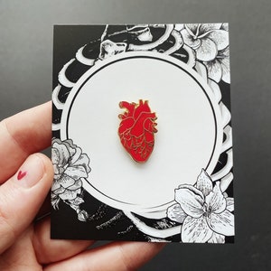 K&ouml;nnte beinhalten: Eine goldfarbene Emaille-Anstecknadel mit einem roten anatomischen Herzen. Die Nadel wird auf einer wei&szlig;en Karte mit schwarz-wei&szlig;en Blumenillustrationen gezeigt.