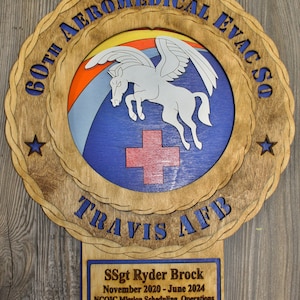 May include: Wooden plaque featuring a circular design with the text "60th Aeromedical Evac Sq" and "Travis AFB". The center depicts a winged horse over a cross. Includes text: SSgt Ryder Brock, dates, and a quote.