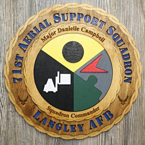 May include: Wooden plaque with the text "71st Aerial Support Squadron" and "Langley AFB" in blue. The center features a segmented design with a globe, a red arrow, and a white forklift. The plaque is a light brown color.