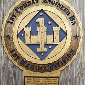 May include: A wooden plaque with a blue and brown emblem of the 1st Combat Engineer Battalion, 1st Marine Division. The emblem features a castle with the number "1" and the words "Guadalcanal" inscribed. The plaque also includes a dedication to 1stLt Matthew T. Thompson, with dates of service and a message of thanks from the Marines and Sailors of 1st CEB.