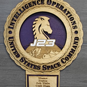 May include: Wooden plaque with the text "UNITED STATES SPACE COMMAND INTELLIGENCE OPERATIONS". The plaque features a horse head silhouette, the letters "J23", and a world map. A smaller plaque below has fields for name, dates, job title, and call sign.