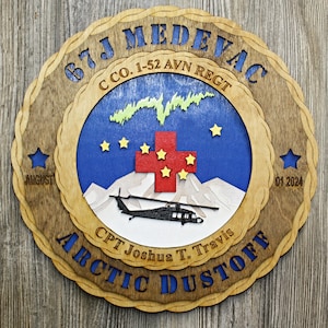 May include: A wooden plaque with a brown background and a blue circle in the center. The circle features a red cross with yellow stars, a black helicopter, and the text "67J MEDEVAC C CO. 1-52 AVN REGT ARCTIC DUSTOFF CPT Joshua T. Travis AUGUST 01 2024".