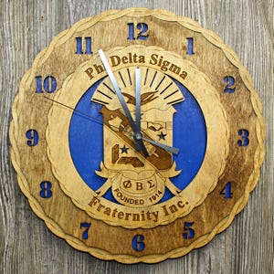 May include: Wooden wall clock with a layered design. The clock features a blue center with the words "Ph Delta Sigma Fraternity Inc." and the founding year 1914. The numbers are blue, and the clock hands are silver.