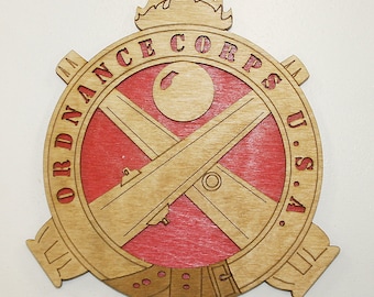 May include: A round wooden plaque with a red background and a brown wood overlay. The plaque features a crossed cannon design with a cannonball and a flame above. The text "ORDNANCE CORPS U.S.A." is written around the edge of the plaque.