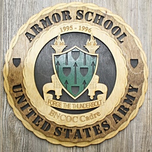 May include: A wooden plaque with a brown and black design. The plaque features the words "Armor School", "United States Army", "BNCOC Cadre", and "Forge the Thunderbolt". The plaque also includes a shield with green and black accents and the years "1995-1996".