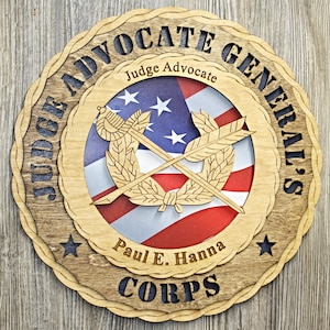 May include: Wooden plaque with the text "JUDGE ADVOCATE GENERAL'S CORPS" and "Judge Advocate" in a circular design. The center features an American flag design with crossed arrows and laurel leaves. The name "Paul E. Hanna" is also included.